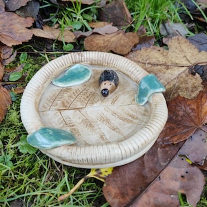 May include: A handmade ceramic bird bath featuring a leaf pattern and a small hedgehog ornament. The bath is beige with green leaf details. The hedgehog has a black body and a brown face. The bird bath is surrounded by leaves and grass.