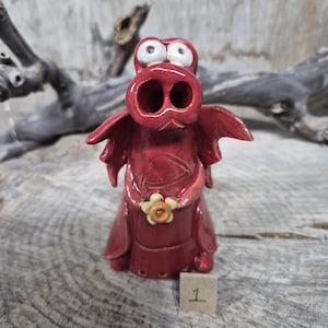 May include: A quirky, red ceramic dragon figurine with large, white-eyed eyes and small wings. The dragon has a flower accent and is sitting on a wooden surface. A small tag with the number 1 is visible.
