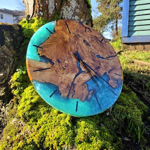 May include: A round wooden clock with a turquoise resin border. The clock face features natural wood grain and black hour markers and hands. The clock is displayed outdoors, resting on a moss-covered tree trunk.