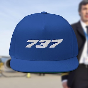 Pode incluir: Boné azul royal de aba reta com o número "737" bordado em branco. O boné tem a parte inferior da aba verde e um botão na parte superior. O fundo está desfocado, mas sugere um tema de aviação.