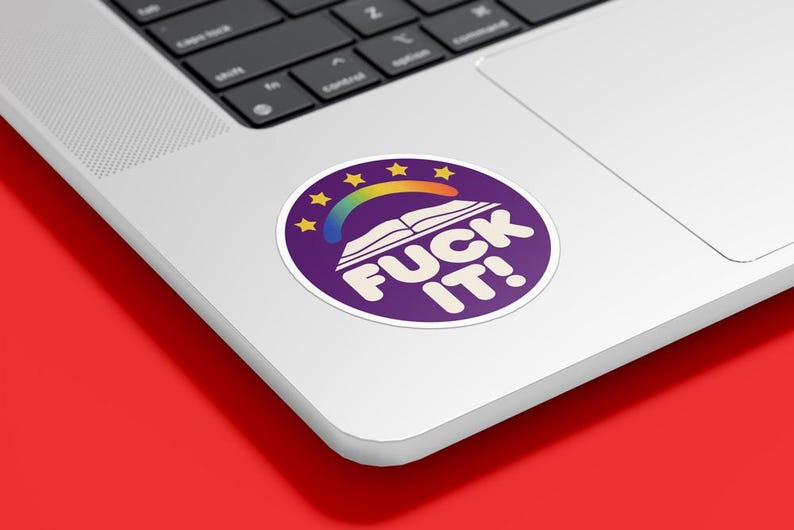 May include: A circular sticker with a purple base, displaying a rainbow, stars, and an open book design. The sticker has the words "FUCK IT!" in white, bold letters. The sticker is placed on a laptop.
