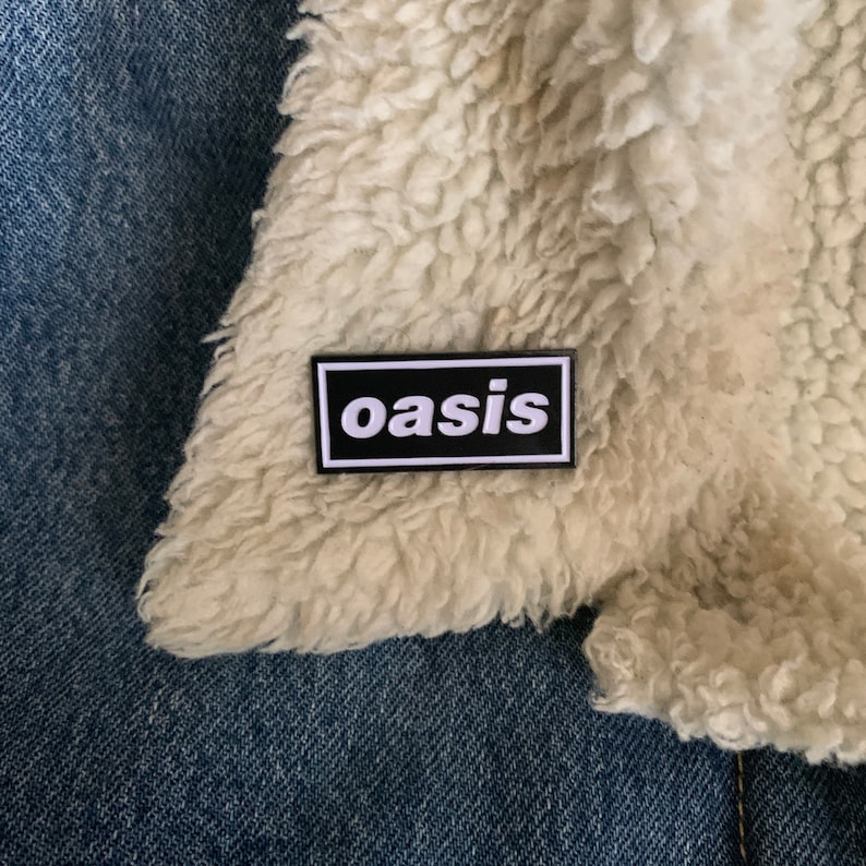 May include: A rectangular black and white patch with the word "oasis" in a white font. The patch is on a cream-colored, fluffy fabric, possibly a jacket lining, set against a blue denim background.