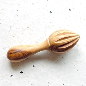 Op de afbeelding: Een houten citruspers met een lichtbruine kleur en een getextureerd oppervlak. De citruspers ligt op een wit gespikkeld oppervlak.