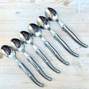 May include: Six silver spoons with curved handles and a polished finish. The spoons are arranged in a row on a light wood surface.