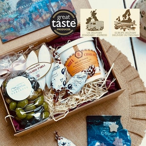 Puede incluir: Una caja de regalo llena de productos gourmet. La caja contiene un tarro de miel, caramelos envueltos, aceitunas y galletas. La caja está etiquetada como "Great Taste Producer". La caja está sobre una superficie de arpillera.