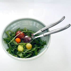 以下が含まれることがあります： 薄い青色のセラミックボウルに、緑の葉物野菜、赤とオレンジのチェリートマト、銀製のサラダサーバー2本が入ったサラダが入っています。
