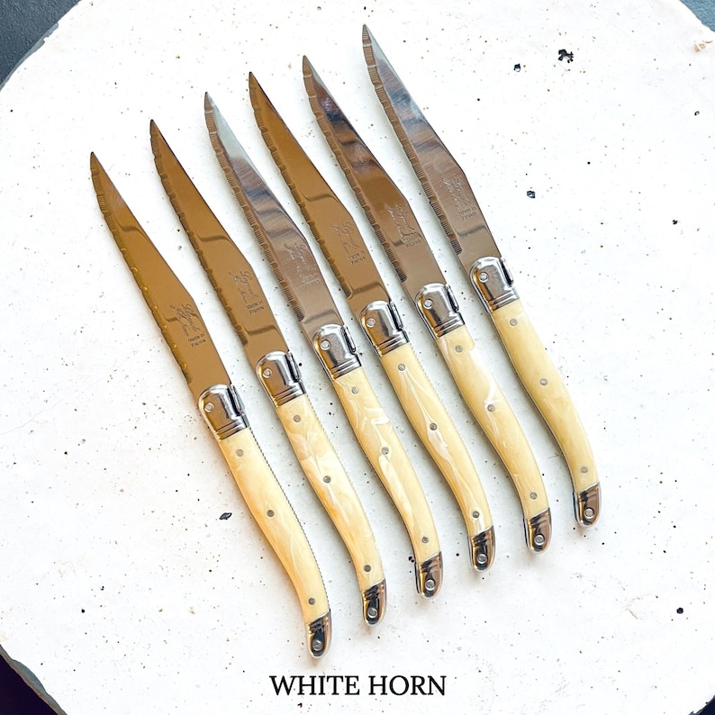 May include: Set of six white horn handled steak knives with stainless steel blades. The knives have a serrated edge and are made in France. The handles are made of white horn and have a natural wood grain pattern.