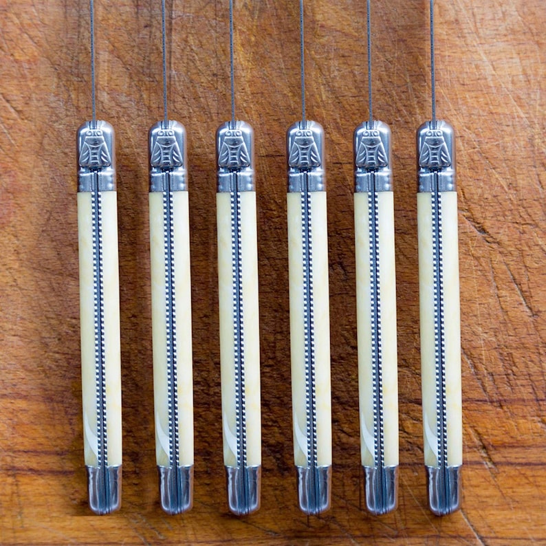 May include: Seven silver pocket knives with ivory-colored handles are lined up on a wooden surface. The knives have a decorative design on the handles.