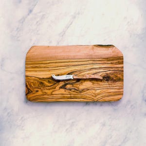 May include: A rectangular wooden cutting board with a cheese knife. The board is made of olive wood, displaying a prominent wood grain pattern. The cheese knife has a curved blade and a wooden handle. The background is a marble surface.