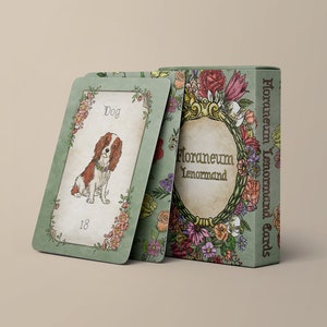 以下が含まれることがあります： カラフルな花々で装飾された緑色の箱に入ったFloraneum Lenormandカードデッキ。1枚のカードが提示されており、茶色と白の犬のイラスト、Dogという文字、数字の18が描かれ、緑色の背景に花柄の縁取りが施されています。