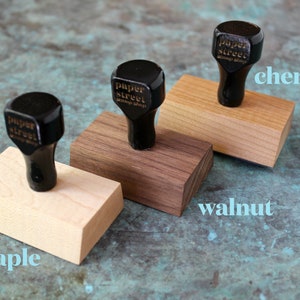 Pu&ograve; includere: Tre blocchi di legno con timbri in gomma nera in cima. I timbri sono etichettati "paper street stamp shop". I blocchi sono etichettati "acero", "noce" e "ciliegio".