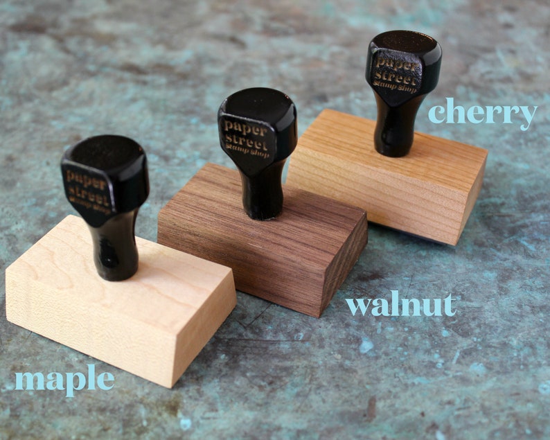 Pu&ograve; includere: Tre blocchi di legno con timbri in gomma nera in cima. I timbri sono etichettati "paper street stamp shop". I blocchi sono etichettati "acero", "noce" e "ciliegio".