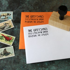May include: A wooden address stamp with a black handle next to an orange and white envelope, both stamped with "THE GREYSONS" and address details. Four vintage wildlife conservation stamps are also visible, featuring various animals and birds.