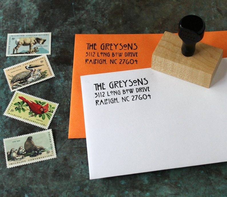 May include: A wooden address stamp with a black handle next to an orange and white envelope, both stamped with "THE GREYSONS" and address details. Four vintage wildlife conservation stamps are also visible, featuring various animals and birds.