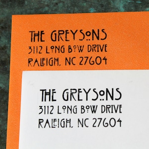 May include: Two sheets of paper, one orange and one white, each with the text "THE GREYSONS" and an address printed in black. The address includes "3112 LONG BOW DRIVE, RALEIGH, NC 27604".