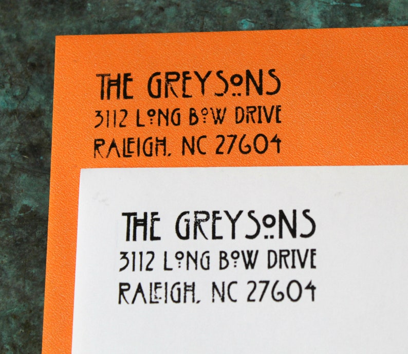 May include: Two sheets of paper, one orange and one white, each with the text "THE GREYSONS" and an address printed in black. The address includes "3112 LONG BOW DRIVE, RALEIGH, NC 27604".