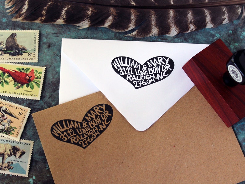 May include: A white envelope with a black heart-shaped stamp that reads "WILLIAM & MARY 3112 LONG BOW DR RALEIGH, NC 27604". The envelope is on a brown surface with other stamps.