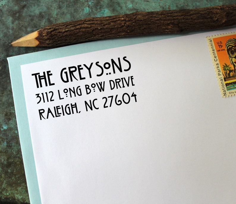 May include: A white envelope with the address "THE GREYSONS, 3112 LONG BOW DRIVE, RALEIGH, NC 27604" in black ink. A wooden pencil and a postage stamp are also visible.