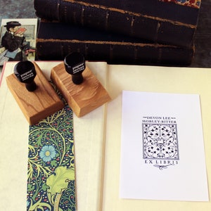 May include: Two wooden stamp blocks with black rubber stamps that read "Paper Street" on a white book with a bookmark. The book is open to a white card with black text that reads "Devon Lee Mobley-Ritter Ex Libris" with a decorative design.