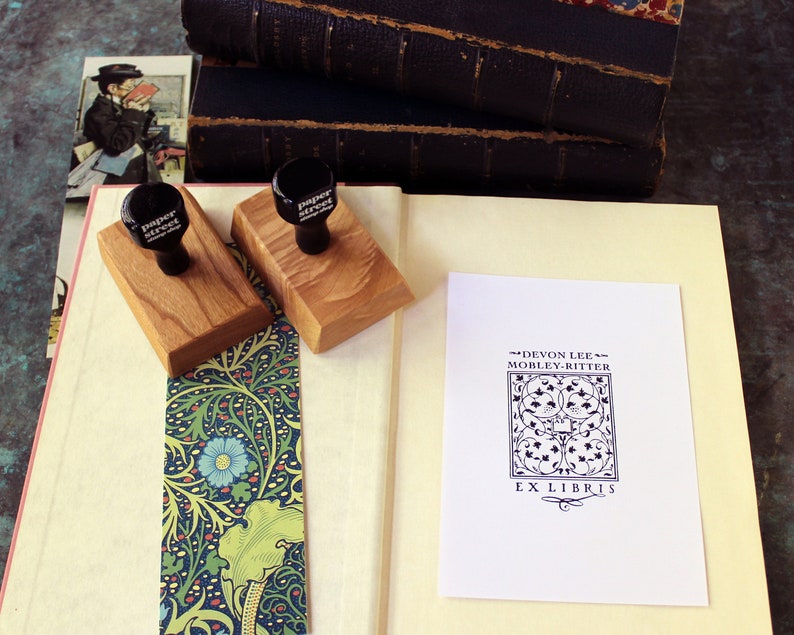May include: Two wooden stamp blocks with black rubber stamps that read "Paper Street" on a white book with a bookmark. The book is open to a white card with black text that reads "Devon Lee Mobley-Ritter Ex Libris" with a decorative design.