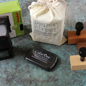 Pu&ograve; includere: Un set di timbri in gomma nera con basi in legno, un tampone d'inchiostro nero e una borsa di tela bianca con il testo "paper street stamp shop" stampato su di essa.