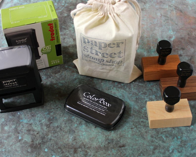 Pu&ograve; includere: Un set di timbri in gomma nera con basi in legno, un tampone d'inchiostro nero e una borsa di tela bianca con il testo "paper street stamp shop" stampato su di essa.