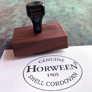 Pu&ograve; includere: Un timbro in legno con una testa di timbro in gomma nera che recita "paper street stamp shop". Il timbro &egrave; su una superficie bianca con un timbro circolare nero che recita "GENUINE HORWEEN 1905 SHELL CORDOVAN" con un simbolo di marchio registrato.