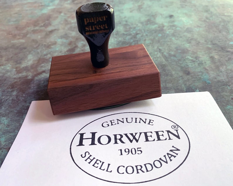 Pu&ograve; includere: Un timbro in legno con una testa di timbro in gomma nera che recita "paper street stamp shop". Il timbro &egrave; su una superficie bianca con un timbro circolare nero che recita "GENUINE HORWEEN 1905 SHELL CORDOVAN" con un simbolo di marchio registrato.