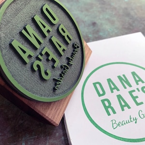 Pu&ograve; includere: Un timbro rotondo in gomma verde con il testo "Dana Rae's Beauty Garden" su una base di legno. Un cartoncino bianco con un cerchio verde e il testo "Dana Rae's Beauty Garden" &egrave; accanto al timbro.