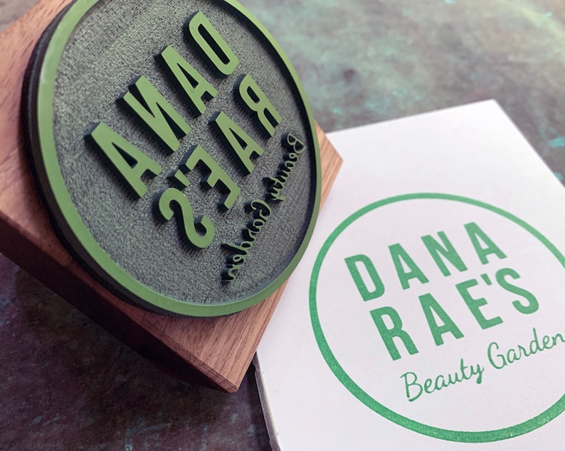 Pu&ograve; includere: Un timbro rotondo in gomma verde con il testo "Dana Rae's Beauty Garden" su una base di legno. Un cartoncino bianco con un cerchio verde e il testo "Dana Rae's Beauty Garden" &egrave; accanto al timbro.