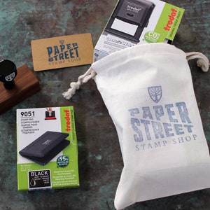 May include: A white canvas bag with a blue logo that says "Paper Street Stamp Shop" and a tree design. The bag is filled with a black ink pad, a wooden stamp, and a business card that says "Paper Street Stamp Shop".