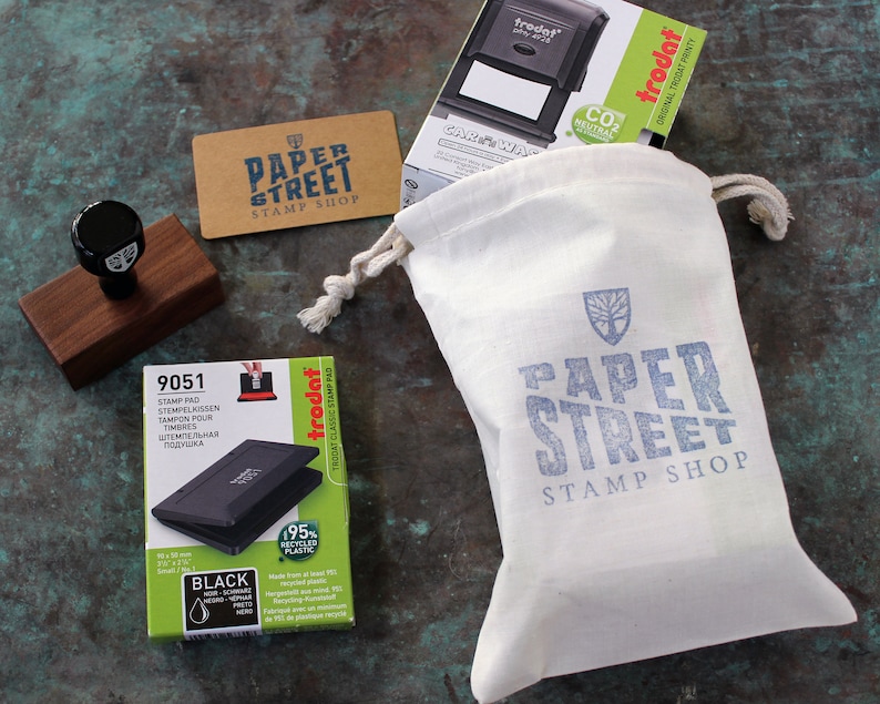 May include: A white canvas bag with a blue logo that says "Paper Street Stamp Shop" and a tree design. The bag is filled with a black ink pad, a wooden stamp, and a business card that says "Paper Street Stamp Shop".