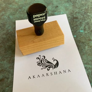 Pu&ograve; includere: Un timbro di legno con un manico nero e il testo "paper street stamp shop" sulla parte superiore. Il timbro &egrave; su un foglio di carta bianco con un disegno di pavone nero e il testo "AKAARSHANA" sotto.