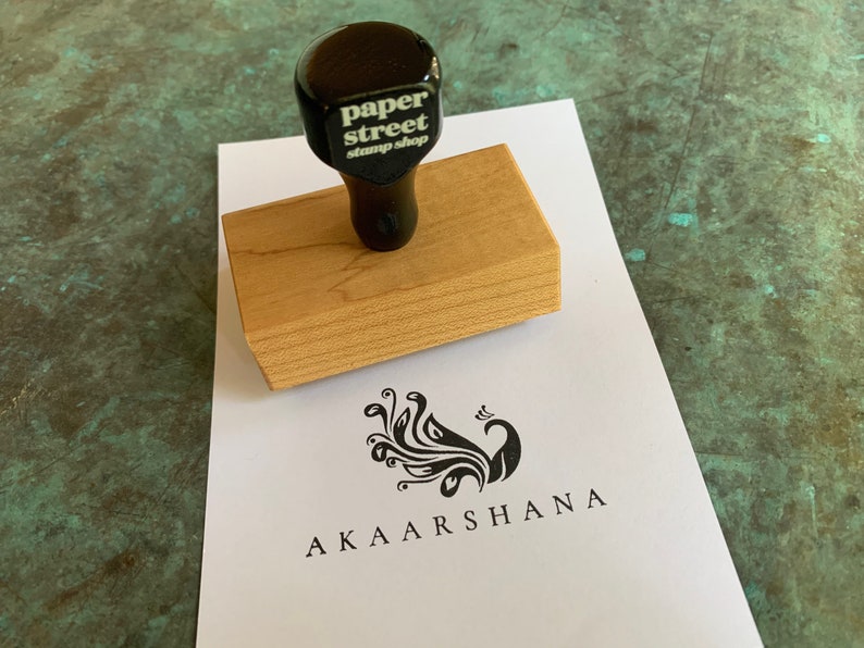 Pu&ograve; includere: Un timbro di legno con un manico nero e il testo "paper street stamp shop" sulla parte superiore. Il timbro &egrave; su un foglio di carta bianco con un disegno di pavone nero e il testo "AKAARSHANA" sotto.