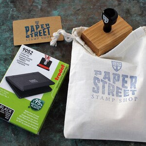 May include: A black and green stamp pad with a white label that says "95% Recycled Plastic" and "trodat" in red. The stamp pad is in a white box with the text "9052" and "STAMP PAD" in black. The box is sitting on a green surface next to a brown card with the text "PAPER STREET STAMP SHOP" in blue. A wooden stamp with a black handle is in a white canvas bag with the text "PAPER STREET STAMP SHOP" in blue.