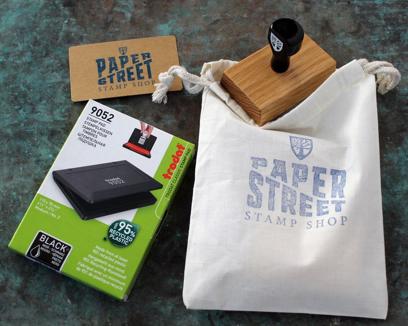May include: A black and green stamp pad with a white label that says "95% Recycled Plastic" and "trodat" in red. The stamp pad is in a white box with the text "9052" and "STAMP PAD" in black. The box is sitting on a green surface next to a brown card with the text "PAPER STREET STAMP SHOP" in blue. A wooden stamp with a black handle is in a white canvas bag with the text "PAPER STREET STAMP SHOP" in blue.