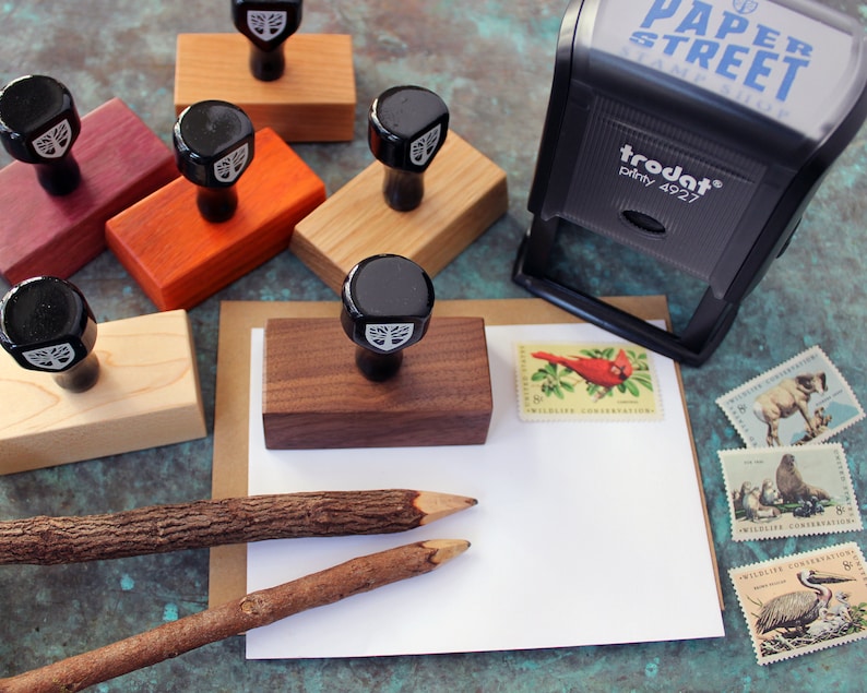 May include: A set of wooden stamps with a tree design, a Trodat stamp with the text "PAPER STREET STAMPS" and the text "trodat&reg; printy 4927", and two wooden pencils on a white card with stamps featuring wildlife conservation.