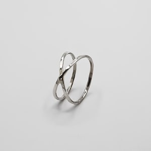 May include: A silver ring featuring an overlapping X design. The ring has a textured surface and is set against a plain white background. The jewellery item is a ring.