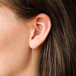 May include: A close-up of an ear with gold bar earrings. The earrings are long, thin, and hang vertically. The person has brown hair. The earrings are made of gold.