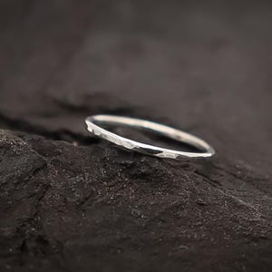 May include: A silver ring with a hammered finish, set against a dark, textured background. The ring is slender and features a faceted design, catching the light. The style is minimalist and refined.