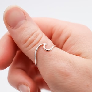 May include: A silver wave ring is displayed on a finger. The ring's design is a minimalist curling ocean wave. The ring is made of a shiny, reflective metal. The design is simple and elegant, suitable for everyday wear.