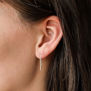 May include: A close-up of an ear with gold-coloured, linear drop earrings. The earrings have a simple, straight bar design. The person has dark brown hair.