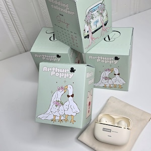 May include: A collection of light green gift boxes with illustrations of a wedding theme. One box features a calendar for July 2025. The boxes are stacked, with a pair of white Lenovo earbuds and a small cloth bag.