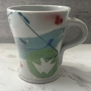 May include: White ceramic mug with a hand-painted abstract design in shades of blue, green, and red. The design features brushstrokes and dots.