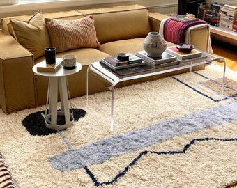 May include: A light brown sectional sofa with a white and gray patterned rug. The sofa has three pillows, two of which are brown and one is a light brown with a white pattern. A clear acrylic coffee table with a curved edge sits in front of the sofa. The coffee table has a few books and a white ceramic vase with a floral design on it. A small white side table with a round top sits next to the sofa.