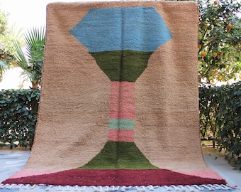 May include: A large, rectangular rug with a geometric design. The rug features a central design in shades of blue, green, and pink against a tan background. The bottom edge has a fringe. The rug is made of a thick, textured material.