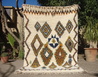 May include: A white Moroccan rug with a geometric pattern in shades of blue, orange, and green. The rug has a fringe border and is made of wool.