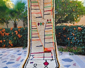 May include: A rectangular, cream-colored rug with a geometric design. The rug features black, orange, green, and red stripes and diamond shapes. The rug is displayed outdoors, leaning against a wall, with a garden in the background.