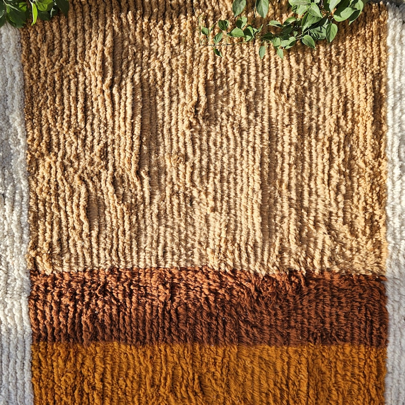 May include: A rectangular, textured rug with a striped design. The rug features a light tan upper section, a dark brown middle section, and a golden-brown lower section. The rug has a white border on the left side. Green leaves are in the upper right corner.