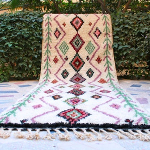 May include: A white, hand-woven rug with a geometric pattern in shades of pink, green, and red. The rug has a fringe border and is laid out on a tiled floor.